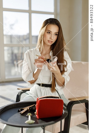 Young woman sitting on a chair and applying make up. Brunette woman sitting in a bright studio with big windows. Girl wearing white blouse and jeans. Young woman sitting on a chair and applying make up. Brunette woman sitting in a bright studio with big windows. Girl wearing white blouse and jeans. 88493522