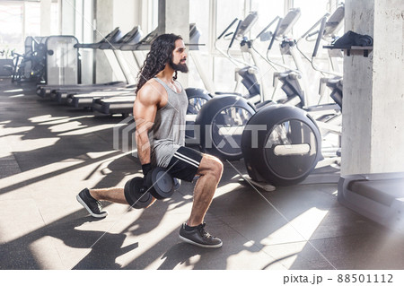 Side view of concentration young adult sportman athlete with long curly hair working out in gym, squating on one knee and holding two dumbbells, doing exercises for legs, squatting. indoor Side view of concentration young adult sportman athlete with long curly hair working out in gym, squating on one knee and holding two dumbbells, doing exercises for legs, squatting. indoor 88501112