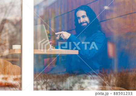 Portrait through the window of cheerful young adult man freelancer in casual style sitting in cafe, talking with friend in laptop, pointing finger to screen, choosing you. outdoor, lifestyle concept 88501233