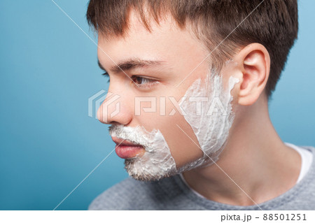 Closeup profile of young confident man looking aside with shaving foam on face, showing smooth soft skin on shaved part of cheek, moisturizing cosmetics. indoor studio shot isolated on blue background 88501251