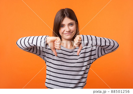 Portrait of dissatisfied young woman with brown hair in long sleeve striped shirt standing, showing disapproval and rejection with thumbs down gesture. indoor studio shot isolated on orange background 88501256