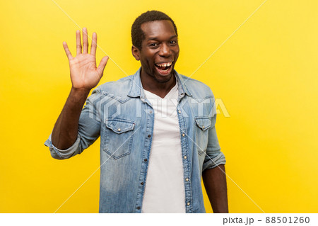 Hello. Portrait of positive handsome man in denim casual shirt with rolled up sleeves smiling friendly and waving hand saying hi, welcoming gesture. indoor studio shot isolated on yellow background 88501260