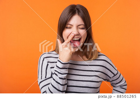 Portrait of funny joyful young woman with brown hair in long sleeve striped shirt standing, picking nose and sticking her tongue out, foolish grimace. indoor studio shot isolated on orange background Portrait of funny joyful young woman with brown hair in long sleeve striped shirt standing, picking nose and sticking her tongue out, foolish grimace. indoor studio shot isolated on orange background 88501265