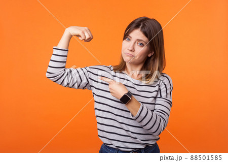 Portrait of proud confident woman with brown hair in long sleeve striped shirt raising arm pointng at biceps, boasting of female power, feminism. indoor studio shot isolated on orange background 88501585