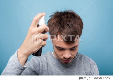 Portrait of young man in casual wear with head down, applying spray to his hair, treating premature hair loss or dandruff, healthcare and treatment. indoor studio shot isolated on blue background Portrait of young man in casual wear with head down, applying spray to his hair, treating premature hair loss or dandruff, healthcare and treatment. indoor studio shot isolated on blue background 88501725