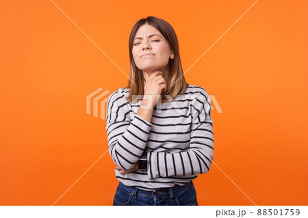 Portrait of ill woman with brown hair in long sleeve striped shirt standing touching neck and frowning from pain, suffering sore throat, flu symptom. indoor studio shot isolated on orange background 88501759