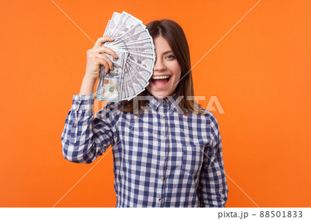 Half face portrait of amazed attractive brunette woman wearing checkered shirt holding dollars and looking at camera with open mouth, lottery winner. indoor studio shot isolated on orange background Half face portrait of amazed attractive brunette woman wearing checkered shirt holding dollars and looking at camera with open mouth, lottery winner. indoor studio shot isolated on orange background 88501833
