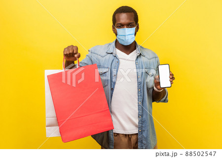 Online shopping and delivery on quarantine. Portrait of young man with medical mask holding packages and showing smartphone and looking with smile. indoor studio shot isolated on yellow background Online shopping and delivery on quarantine. Portrait of young man with medical mask holding packages and showing smartphone and looking with smile. indoor studio shot isolated on yellow background 88502547