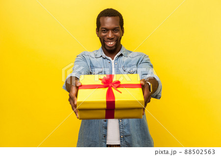 This present for you. Portrait of happy generous man in denim casual shirt giving wrapped gift box and smiling at camera, congratulating on birthday. indoor studio shot isolated on yellow background 88502563