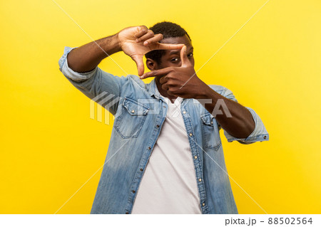 Portrait of focused curious man in denim casual shirt with rolled up sleeves looking through photo frame made of hands, viewing distant with interest. indoor studio shot isolated on yellow background 88502564