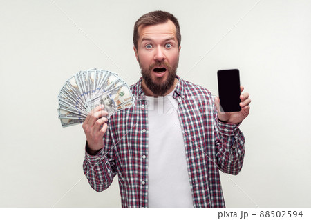 Mobile banking app. Portrait of amazed bearded man in casual plaid shirt holding smartphone and lot of money, looking at the camera in surprise shock. indoor studio shot isolated on white background Mobile banking app. Portrait of amazed bearded man in casual plaid shirt holding smartphone and lot of money, looking at the camera in surprise shock. indoor studio shot isolated on white background 88502594