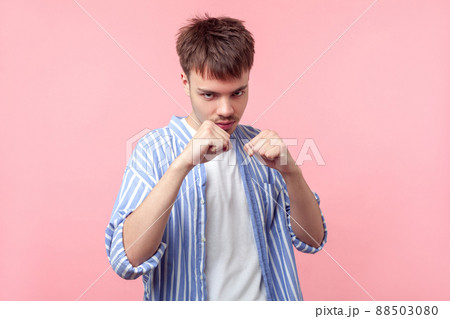 Portrait of angry brown-haired man with small beard and mustache in casual striped shirt standing with clenched fists and furious look, ready to punch. indoor studio shot isolated on pink background Portrait of angry brown-haired man with small beard and mustache in casual striped shirt standing with clenched fists and furious look, ready to punch. indoor studio shot isolated on pink background 88503080