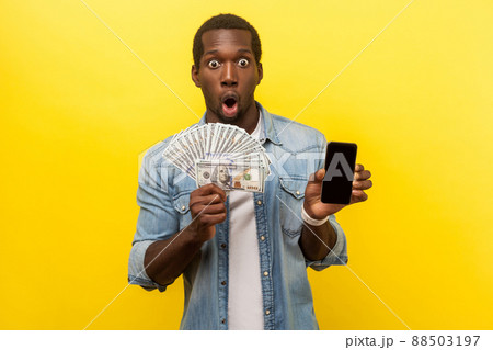 Wow, profitable service, online banking. Portrait of surprised excited man in denim shirt holding dollars and smartphone, making money in mobile application. studio shot isolated on yellow background 88503197