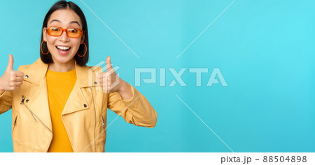 Enthusiastic korean girl looks excited and shows thumbs up, approves smth awesome, excellent choice and quality, stands over blue background 88504898