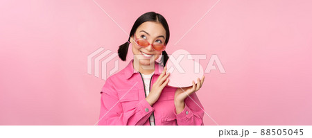 Image of stylish asian girlfriend in sunglasses, guessing whats inside gift box, heart-shaped present, standing over pink background Image of stylish asian girlfriend in sunglasses, guessing whats inside gift box, heart-shaped present, standing over pink background 88505045
