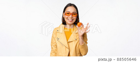 Beautiful asian girl in sunglasses showing tangerine and smiling, looking happy, posing in yellow against studio background 88505646