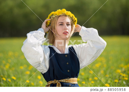 Young woman in national clothes wearing yellow dandelion wreath in spring field. Springtime Young woman in national clothes wearing yellow dandelion wreath in spring field. Springtime 88505958