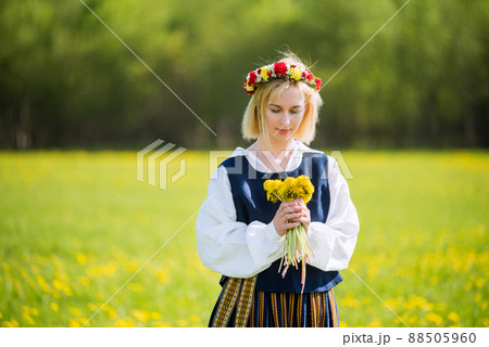 Young woman in national clothes wearing yellow dandelion wreath in spring field. Springtime Young woman in national clothes wearing yellow dandelion wreath in spring field. Springtime 88505960