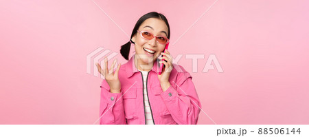Beautiful korean female model in sunglasses, talking on mobile phone with happy face, using cellular service to call friend on smartphone, standing over pink background 88506144