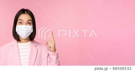Close up portrait of businesswoman, asian lady in office suit and medical face mask, looking and pointing up, showing company logo or banner on top, pink background 88506532