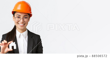 Close-up of happy asian female engingeer, real estate agent in helmet and business suit holding house miniature and smiling, architect working over renovation project, white background 88506572