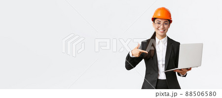 Smiling pleased asian female chief engineer showing chart with enterprise profit, making presentation during investors meeting, wearing safety helmet, pointing at laptop screen, white background 88506580