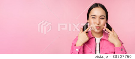 Close up portrait of young asian girl showing her dimples, poking cheeks silly and making funny faces, standing over pink background 88507760