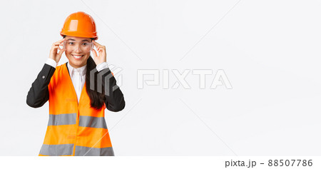 Smiling pretty female asian engineer, construction manager in safety helmet and glasses following protocol before enterting building area or enterprise, standing white background 88507786