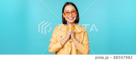 Portrait of asian woman in sunglasses, looking hopeful, flattered, smiling happy, standing over blue background 88507982