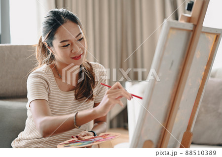 Positive young woman holding paintbrush and palette painting on canvas. 88510389
