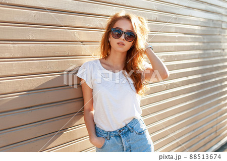 Stylish beautiful young model woman with sunglasses in a white T-shirt and blue vintage jeans near a wooden wall at sunset. Fashion hipster summer season 88513674