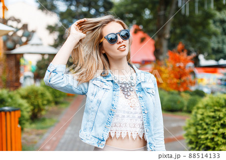 Wonderful young hipster woman in a stylish blue denim jacket in black sunglasses in a vintage lace blouse with a necklace in an amusement park on a sunny spring day. Charming stylish girl travels. Wonderful young hipster woman in a stylish blue denim jacket in black sunglasses in a vintage lace blouse with a necklace in an amusement park on a sunny spring day. Charming stylish girl travels. 88514133