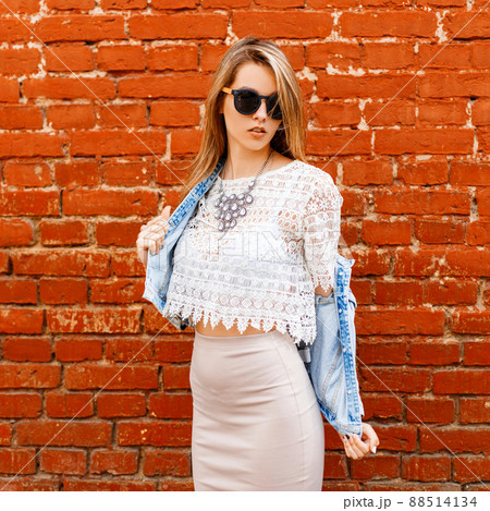 Beautiful sexy young hipster woman in a white lace blouse and denim jacket in black sunglasses in a beige skirt poses near a red brick wall. European modern girl. Beautiful sexy young hipster woman in a white lace blouse and denim jacket in black sunglasses in a beige skirt poses near a red brick wall. European modern girl. 88514134