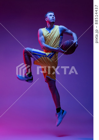 Studio shot of muscled man, basketball player training with ball isolated on purple background in neon light. Goals, sport, motion, activity concepts. 88514437