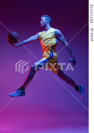Studio shot of muscled man, basketball player training with ball isolated on purple background in neon light. Goals, sport, motion, activity concepts. 88514438