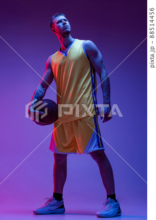 Studio shot of muscled man, basketball player standing with ball isolated on purple background in neon light. Goals, sport, motion, activity concepts. 88514456