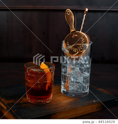 Whiskey with a slice of orange in a crystal glass is on the table in the bar next to the glass with ice and professional tool for making a cocktail. Spiced strong drink. Whiskey with a slice of orange in a crystal glass is on the table in the bar next to the glass with ice and professional tool for making a cocktail. Spiced strong drink. 88516020