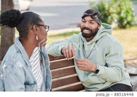 Smiling handsome bearded man in hoodie sitting on bench and telling funny story to black girl in city 88516448