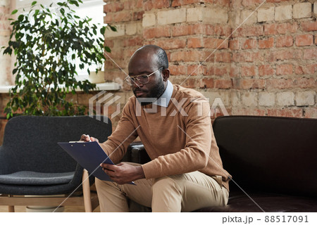 Serious busy young African-American psychologist in eyeglasses sitting on sofa and analyzing notes in clipboard 88517091