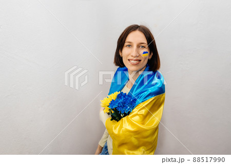 A young girl is wearing the national flag of Ukraine and holding flowers on a white background. The girl has a blue and yellow flag on her cheek. Independence Day of Ukraine. Copy Space. 88517990