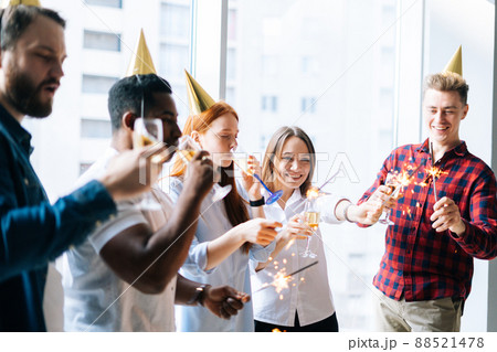 Successful business team of young multi-ethnic businesspeople holding Bengal lights and clinking glasses of champagne at corporate party, on background of window and cityscape with skyscrapers. Successful business team of young multi-ethnic businesspeople holding Bengal lights and clinking glasses of champagne at corporate party, on background of window and cityscape with skyscrapers. 88521478