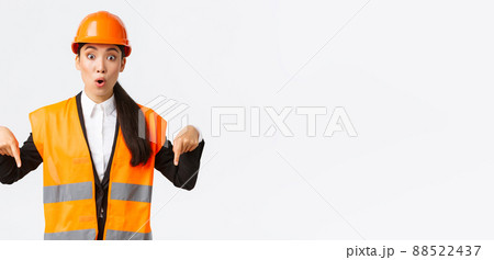 Amazed and interested asian female architect, industrial woman in reflective jacket, safety helmet pointing fingers down and looking impressed, talking about building construction, white background Amazed and interested asian female architect, industrial woman in reflective jacket, safety helmet pointing fingers down and looking impressed, talking about building construction, white background 88522437