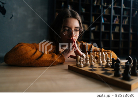 Pensive attractive young woman in elegant eyeglasses thinking about chess move sitting on floor in dark library room, selective focus. Pretty intelligent lady playing logical board game alone at home. 88523030