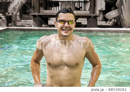 Swimmer. Portrait of smiling athletic man with bare torso wearing glasses for swimming in pool, looking into the camera. Concept of sports, a healthy lifestyle. Sports and fitness cardio exercises. Swimmer. Portrait of smiling athletic man with bare torso wearing glasses for swimming in pool, looking into the camera. Concept of sports, a healthy lifestyle. Sports and fitness cardio exercises. 88539261