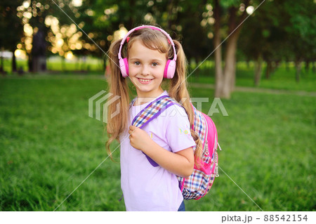 the child is a cute little girl with a school plaid backpack listening to music in pink headphones and smiling against the background of the park 88542154
