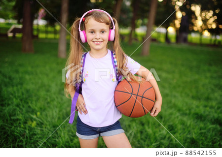 a cute little girl in pink headphones and with a basketball is smiling against the background of greenery and a park. 88542155