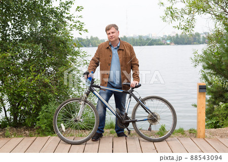 An adult blond man in a brown corduroy jacket is standing by the river in the park with a bicycle 88543094