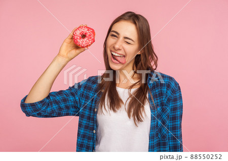 Confectionery and junk food. Portrait of joyous playful girl in checkered shirt sticking out tongue and holding doughnut, having fun with dessert. indoor studio shot isolated on pink background 88550252