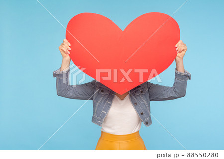 Anonymous woman in casual outfit hiding face behind huge red heart, concept of secret love fondness, unknown lover and affair, romance on Valentines day. indoor studio shot isolated on blue background Anonymous woman in casual outfit hiding face behind huge red heart, concept of secret love fondness, unknown lover and affair, romance on Valentines day. indoor studio shot isolated on blue background 88550280