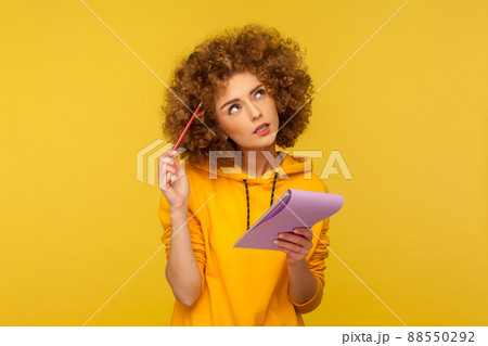 Beautiful pensive woman with Afro hairstyle holding notepad and pencil in hands thinking over plans for startup, wearing casual style hoodie. Indoor studio shot isolated on yellow background. 88550292